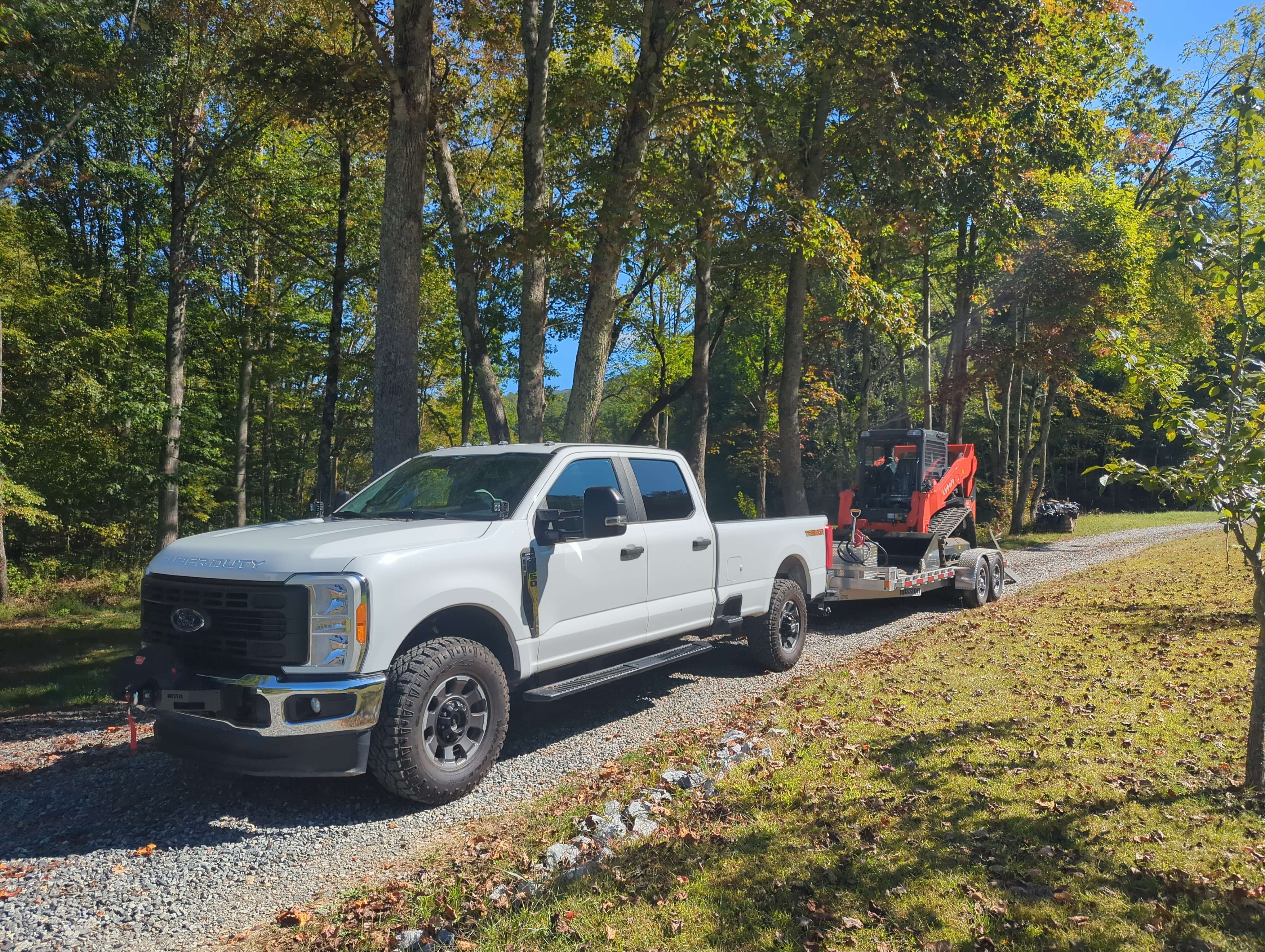 White Ford truck pulling red skid steer on trailer through wooded area
